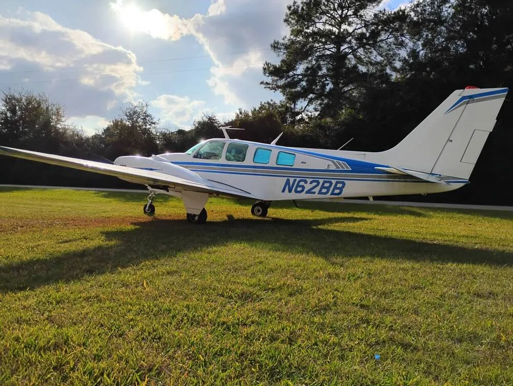 A white Beechcraft Baron N62BB with blue stripes is parked on a grassy field under a partly cloudy sky, viewed from the front-right.