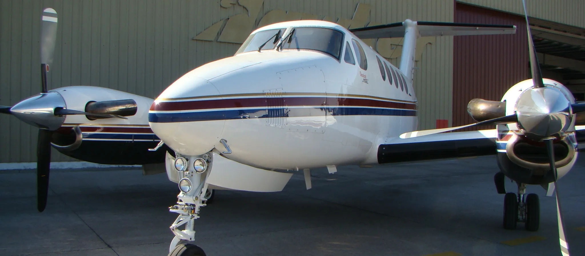 A white twin-engine turboprop aircraft with blue, red, and gold stripes is parked on a tarmac in front of a hangar.