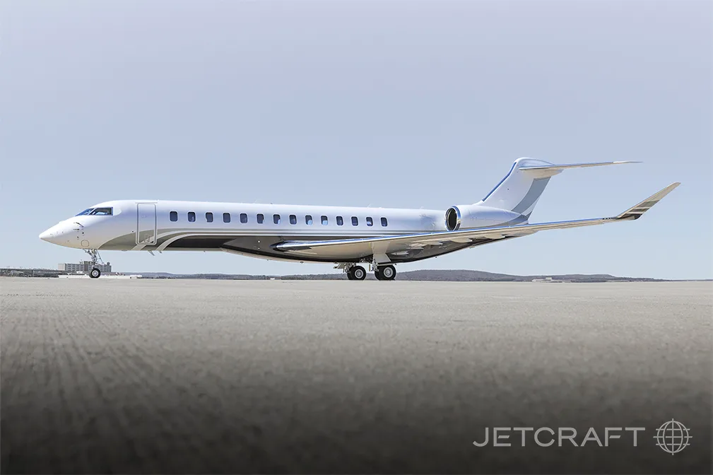 A white and grey Bombardier Global 7500 business jet parked on an airfield under a clear sky.