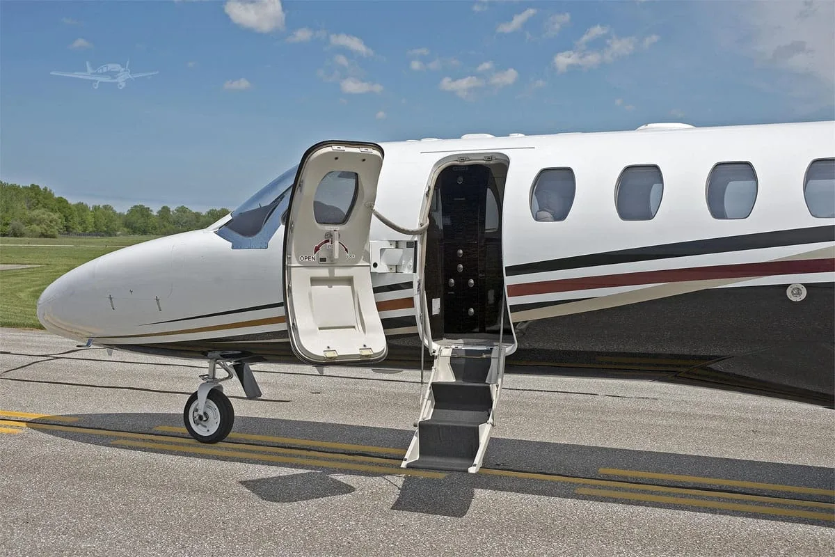 The front section of a white and black business jet with its main entry door open and stairs extended on a sunny tarmac.