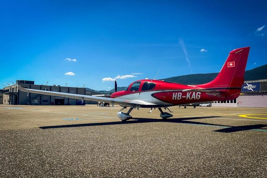 Side view of a red and silver Cirrus SR22 aircraft parked on an airfield with hangars and mountains in the distance.