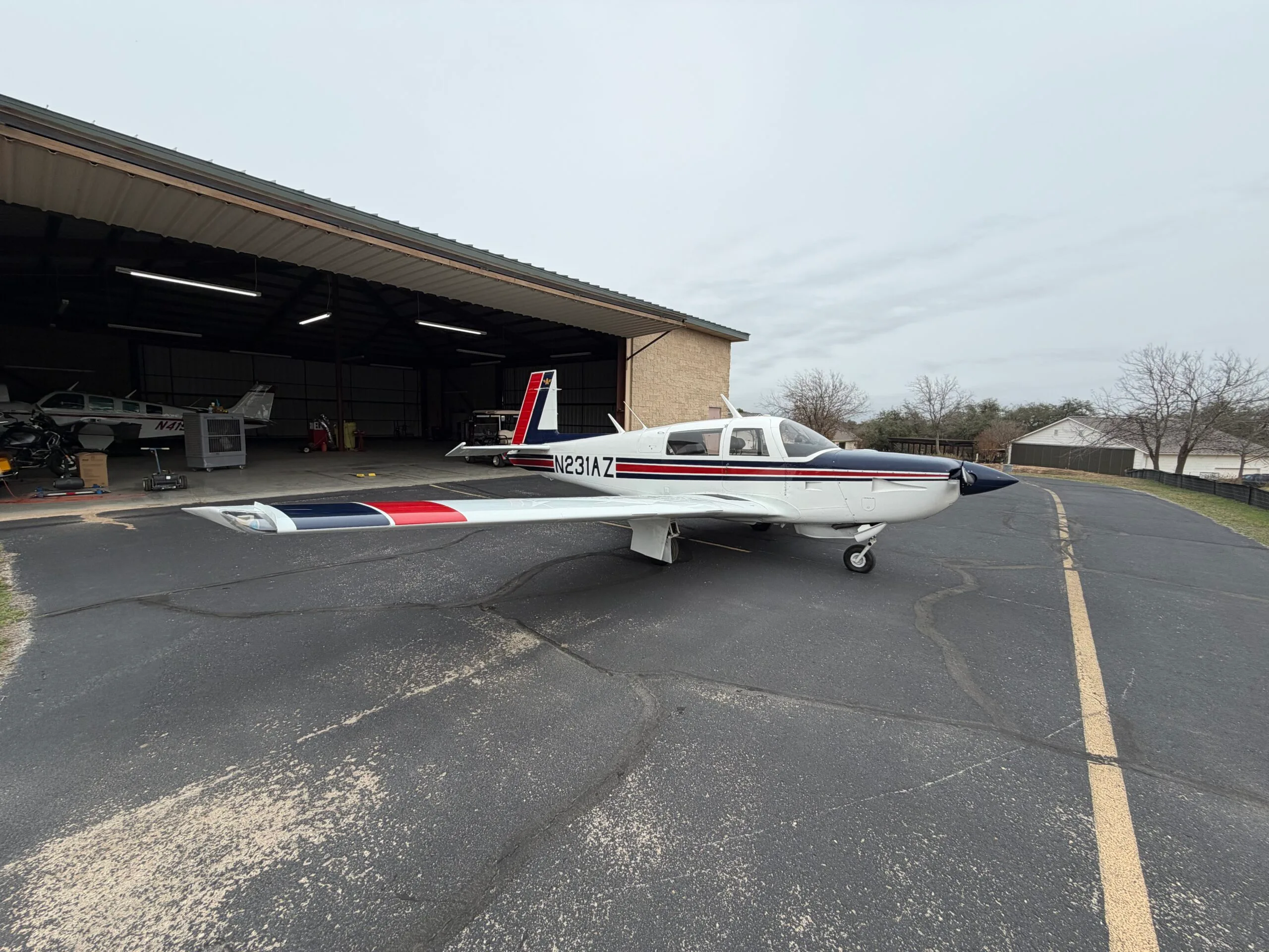 A white single-engine aircraft with blue and red stripes is parked on an asphalt ramp outside an open hangar under an overcast sky.