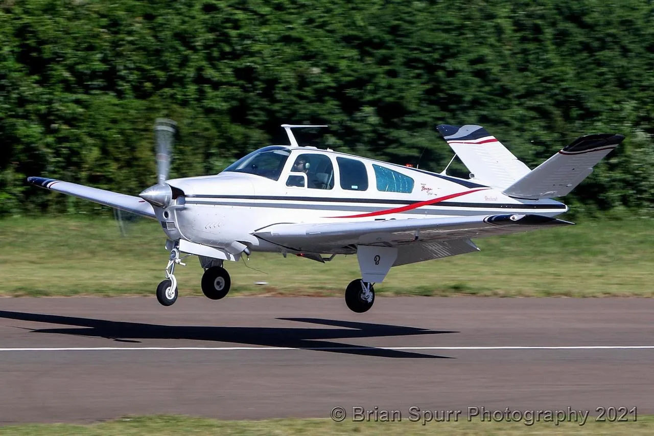 A white Beechcraft Bonanza with red and black stripes is captured just above a runway during takeoff or landing.