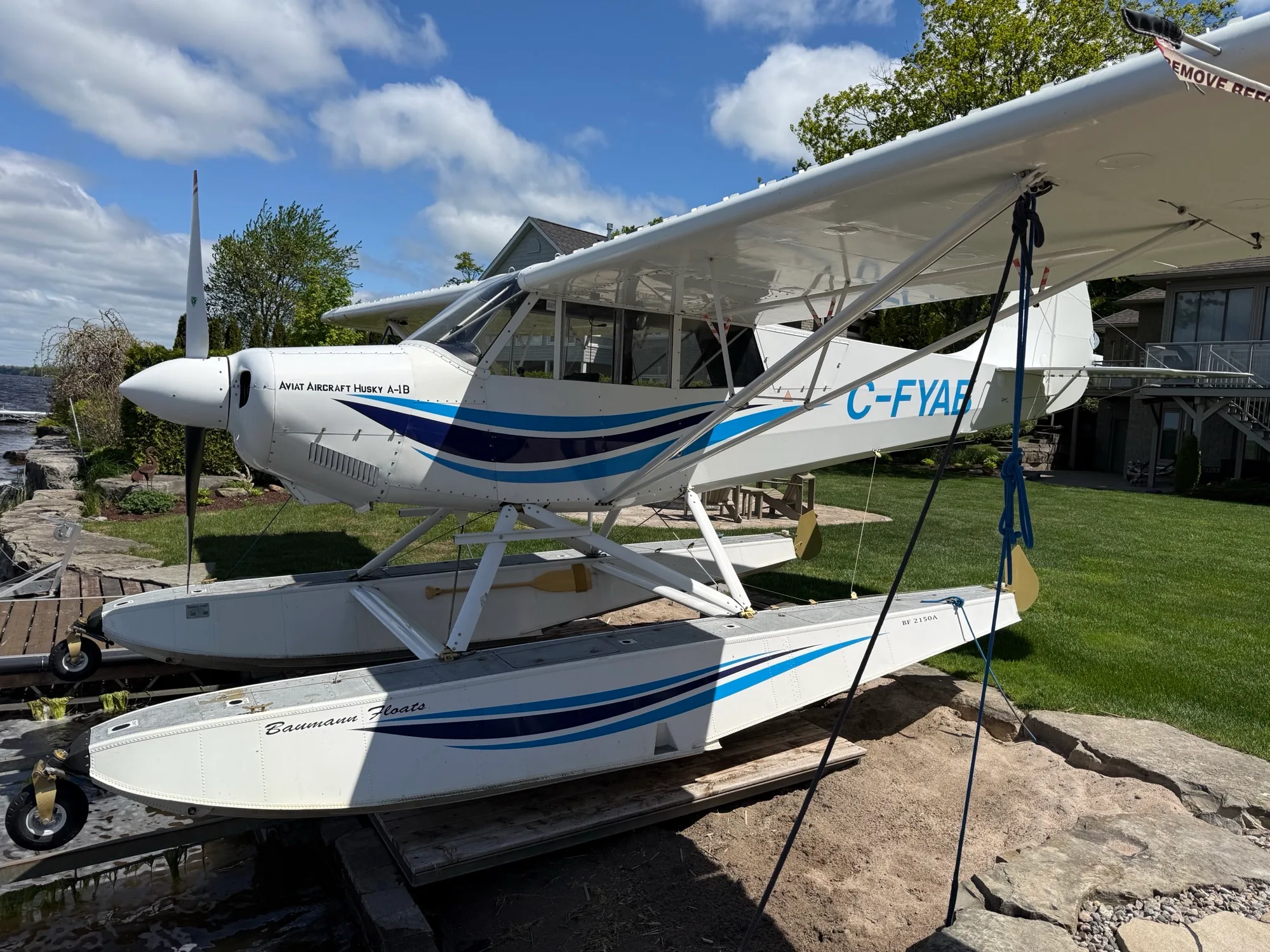 A white Aviat Aircraft Husky A-1B floatplane with blue stripes, parked on a ramp next to a lake on a sunny day.
