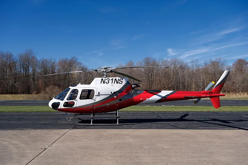A white and red helicopter with registration N31NS is parked on an asphalt surface with trees in the background under a clear blue sky.