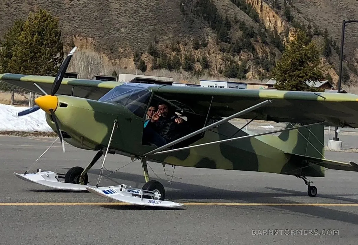 A camouflage-painted light aircraft with skis attached to its wheels has two people visible in the cockpit, parked on an asphalt surface near a mountain.