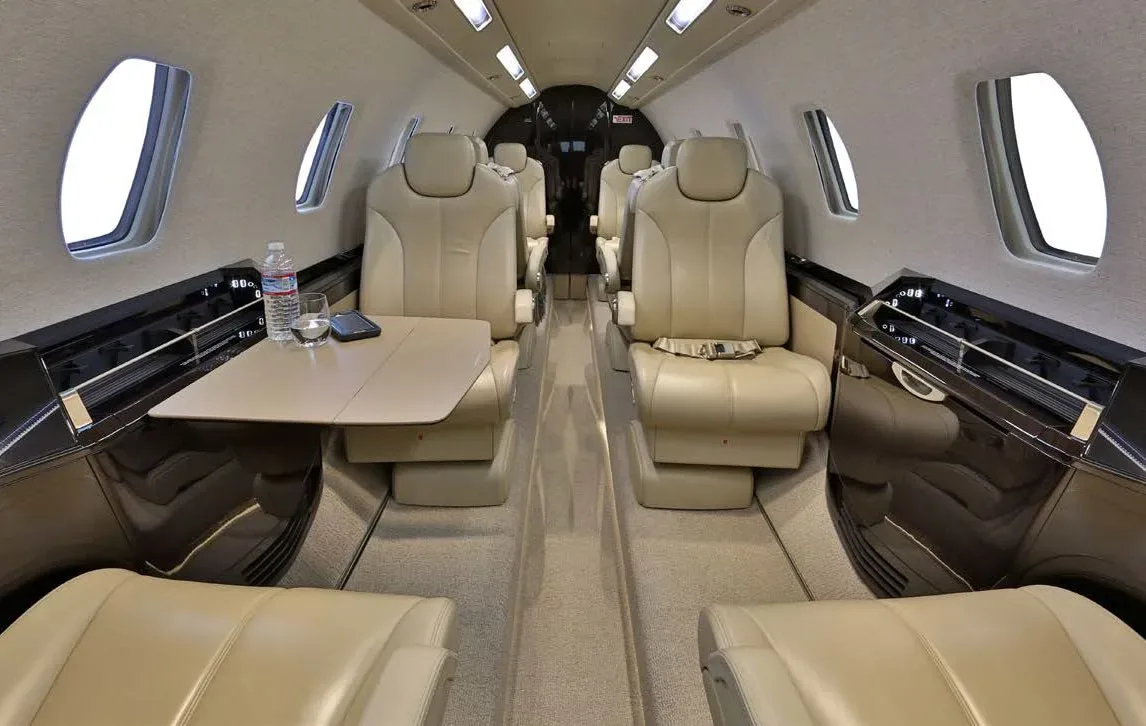 The elegant cabin interior of a business jet, showing tan leather seats, a deployed table with a water bottle, and a phone.