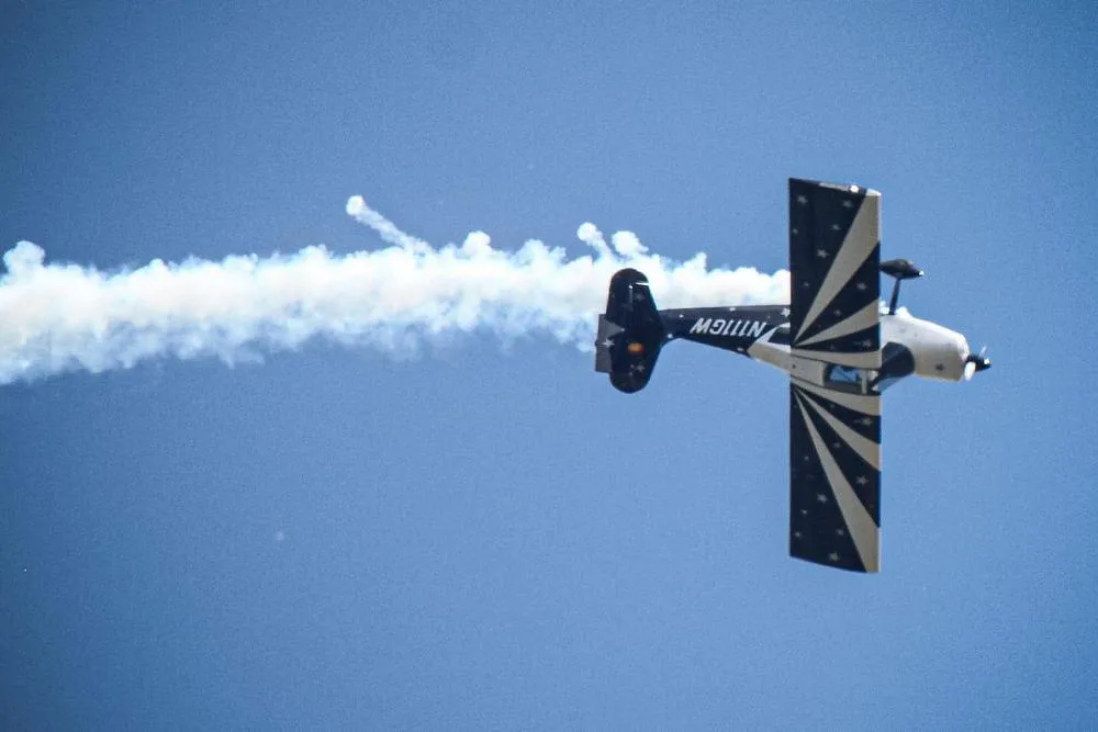 A black and white biplane with a starburst paint scheme, registered N111GW, performing an aerial maneuver with a smoke trail.