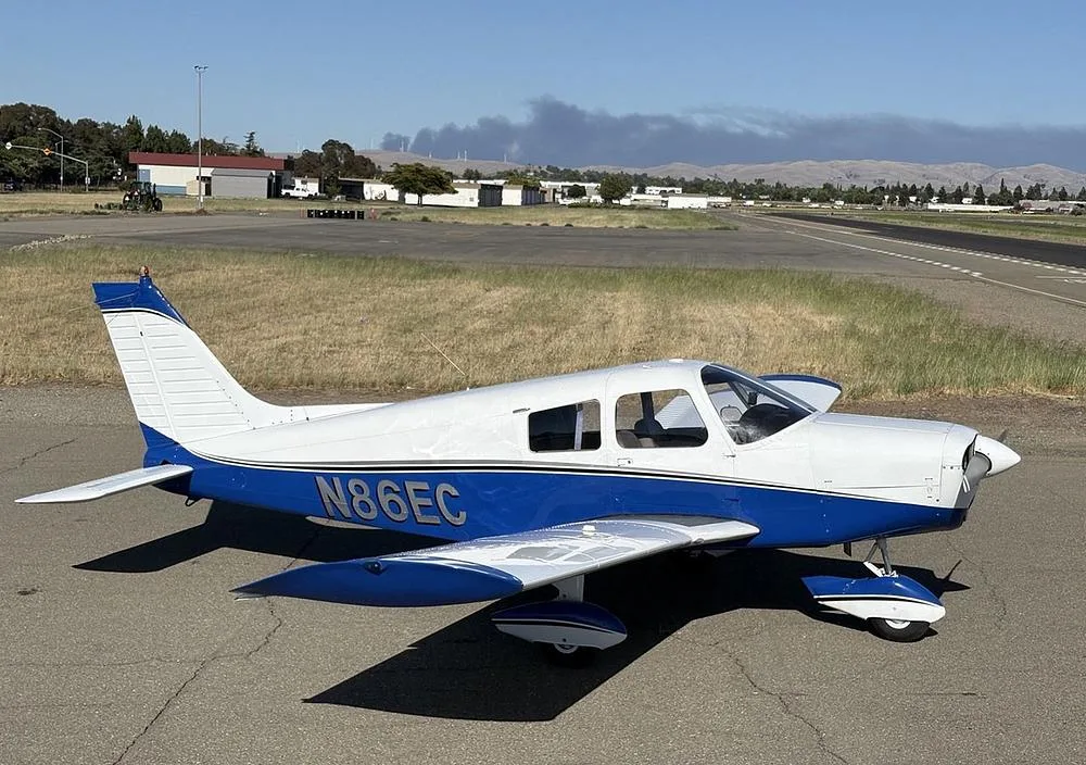 A white and blue Piper Cherokee aircraft (N86EC) parked on an airfield with smoke visible in the distant sky.