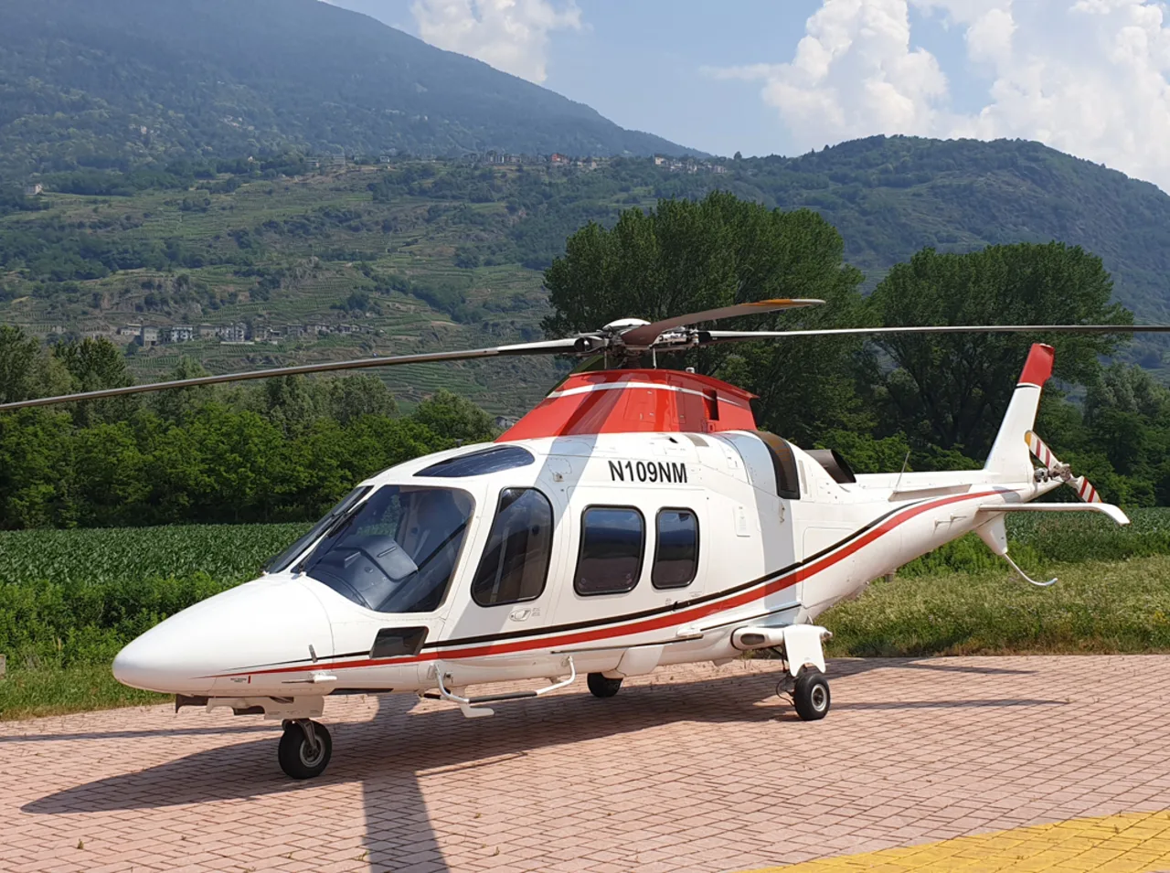 A white helicopter with red and black stripes, registration N109NM, is parked on a paved surface with mountains and green trees in the background.
