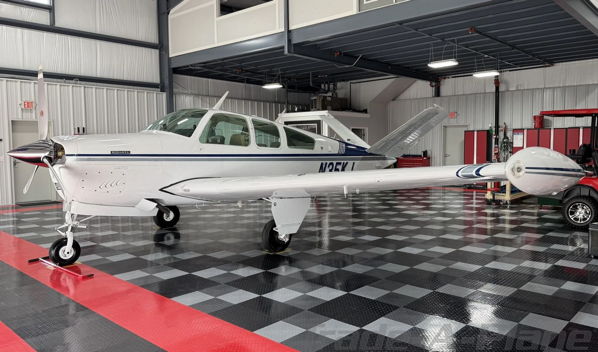 A white Beechcraft Bonanza with blue and grey stripes parked in a hangar with a checkered floor.