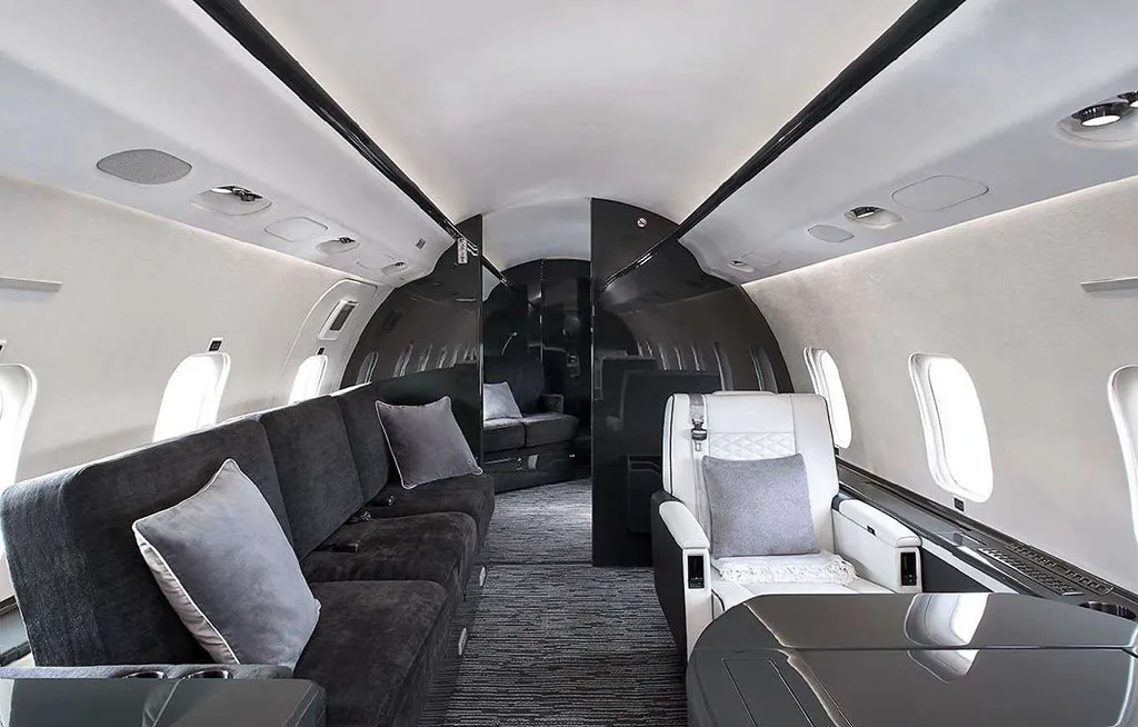 The spacious interior cabin of a private jet, showing a dark grey sofa on the left and a white quilted seat on the right.
