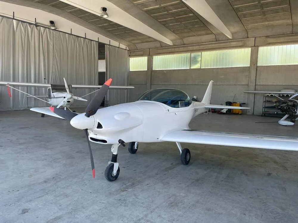 A white Fascination ultralight aircraft with a clear canopy is parked inside a hangar, viewed from the front-left.