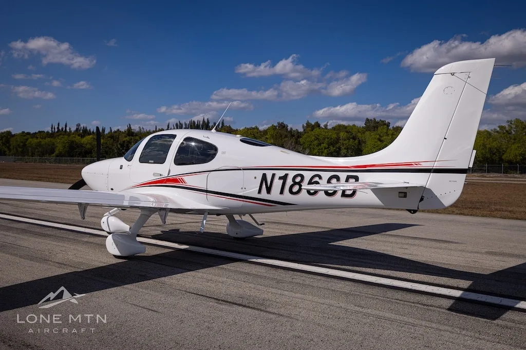 A rear-side view of the white N186SB single-engine aircraft with red and black accent stripes, parked on an airfield.