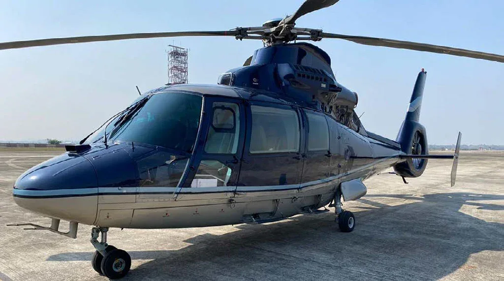 A front-side view of a dark blue and silver Airbus H155 helicopter parked on an airfield with a tower in the distance.