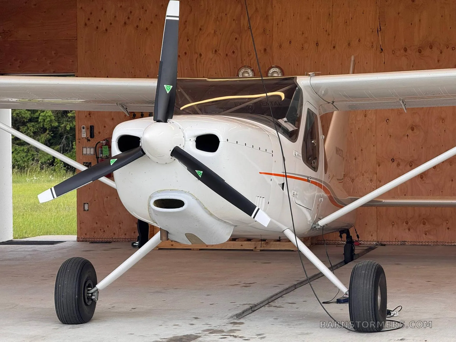 A white single-engine propeller aircraft with orange stripes is parked in a wooden hangar, viewed from the front.