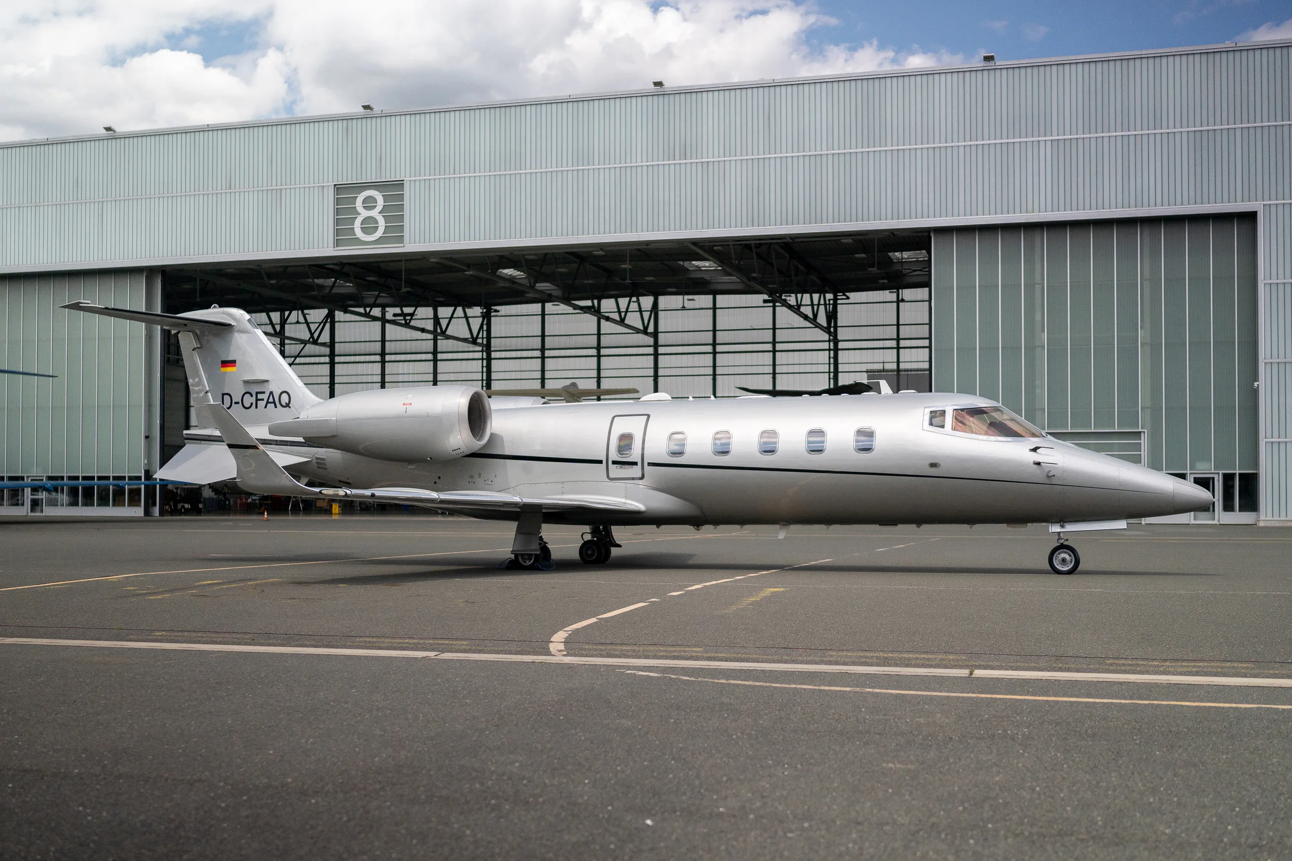 A silver Learjet 60 business jet with registration D-CFAQ parked on a tarmac in front of a hangar.
