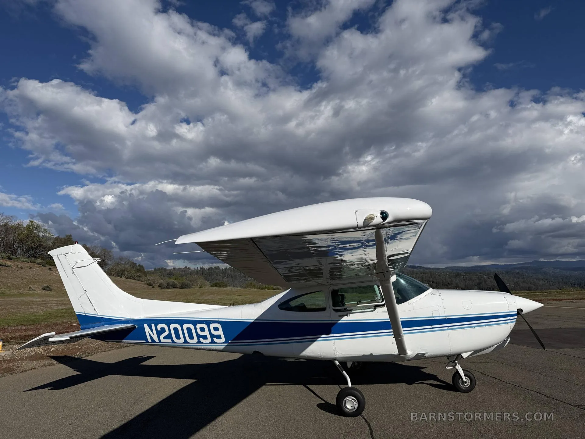 A white Cessna 182 with blue stripes, registration N20099, parked on an asphalt ramp under a dramatic cloudy sky.