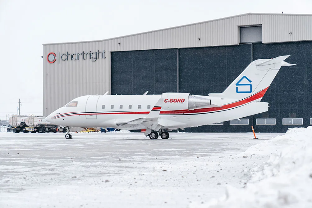 A white business jet with red and blue stripes and a house logo on the tail, parked on a snowy tarmac in front of a Chartright hangar.