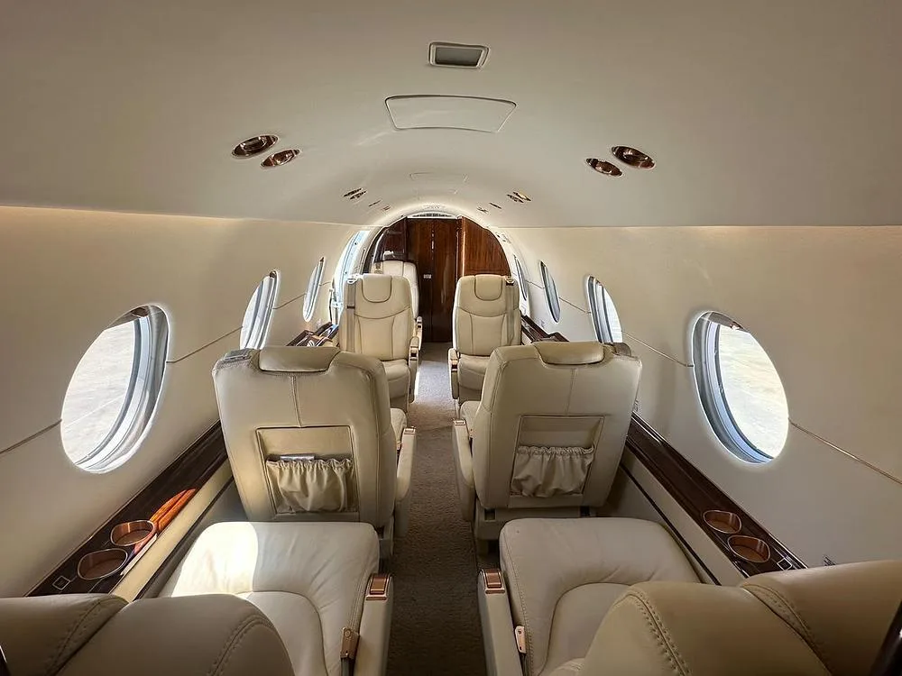 The luxurious interior cabin of a business jet, featuring beige leather seats and large oval windows.