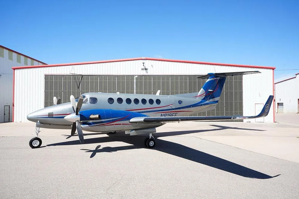 A silver and blue twin-turboprop aircraft, N819EE, parked on an tarmac in front of hangars.