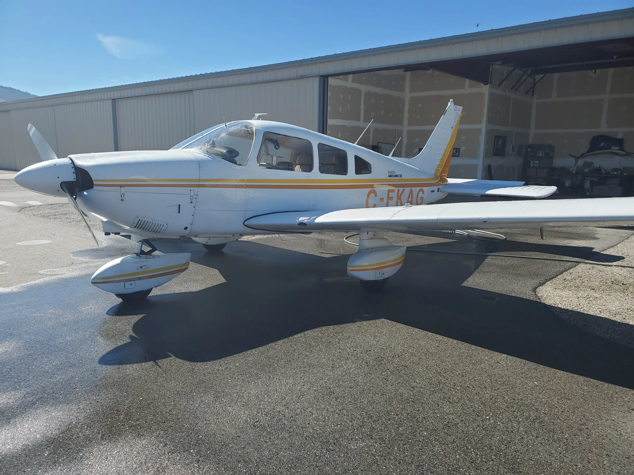 A white Piper Archer II with orange stripes is parked on a wet tarmac outside a hangar on a sunny day.