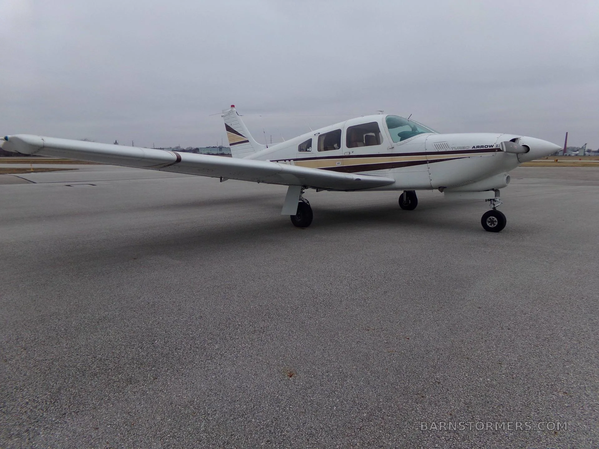 A white Piper Turbo Arrow III aircraft with brown and gold stripes is parked on an asphalt tarmac under an overcast sky, viewed from the front-right.