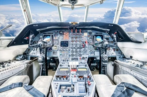 The cockpit of a business jet, showing a complex instrument panel with analog gauges and digital displays, with clouds visible through the windshield.