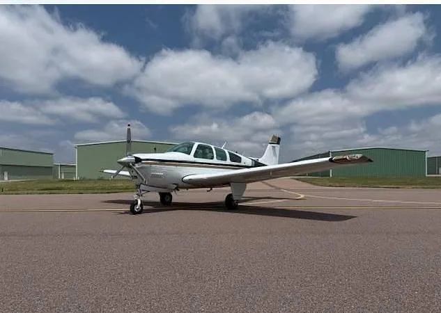 A white and gold Beechcraft Bonanza aircraft is parked on an airfield with green hangars under a cloudy sky.