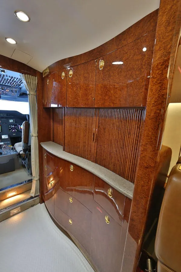 A luxurious aircraft interior shows polished wood cabinetry and a glimpse of the cockpit through an open doorway.