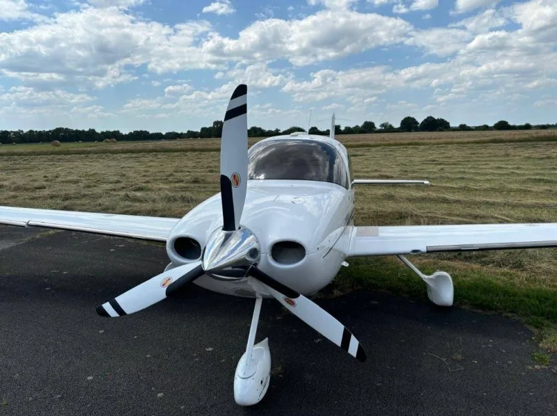 Front view of a white Cirrus SR22 aircraft with a three-bladed propeller on a grassy airfield.