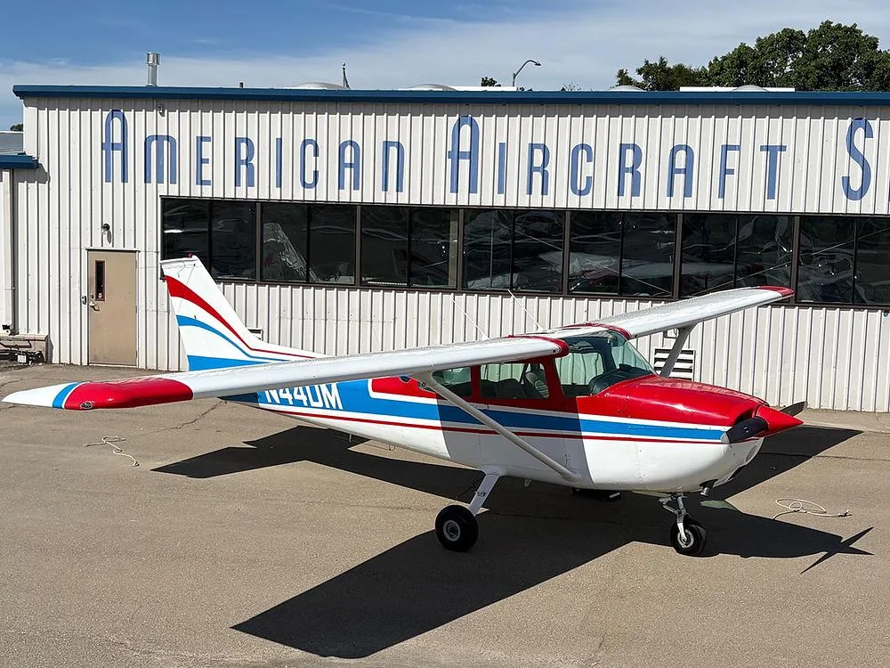 A white Cessna 172 with red and blue stripes, registration N44DM, parked on an asphalt ramp with a building in the background.
