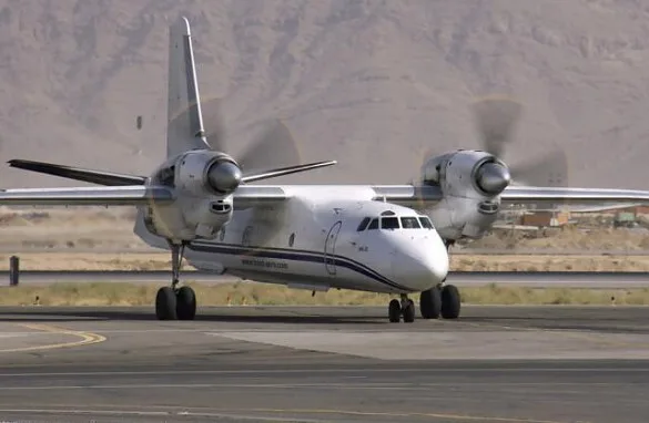 A white An-32 turboprop aircraft with blue stripes on a runway with mountains in the background.