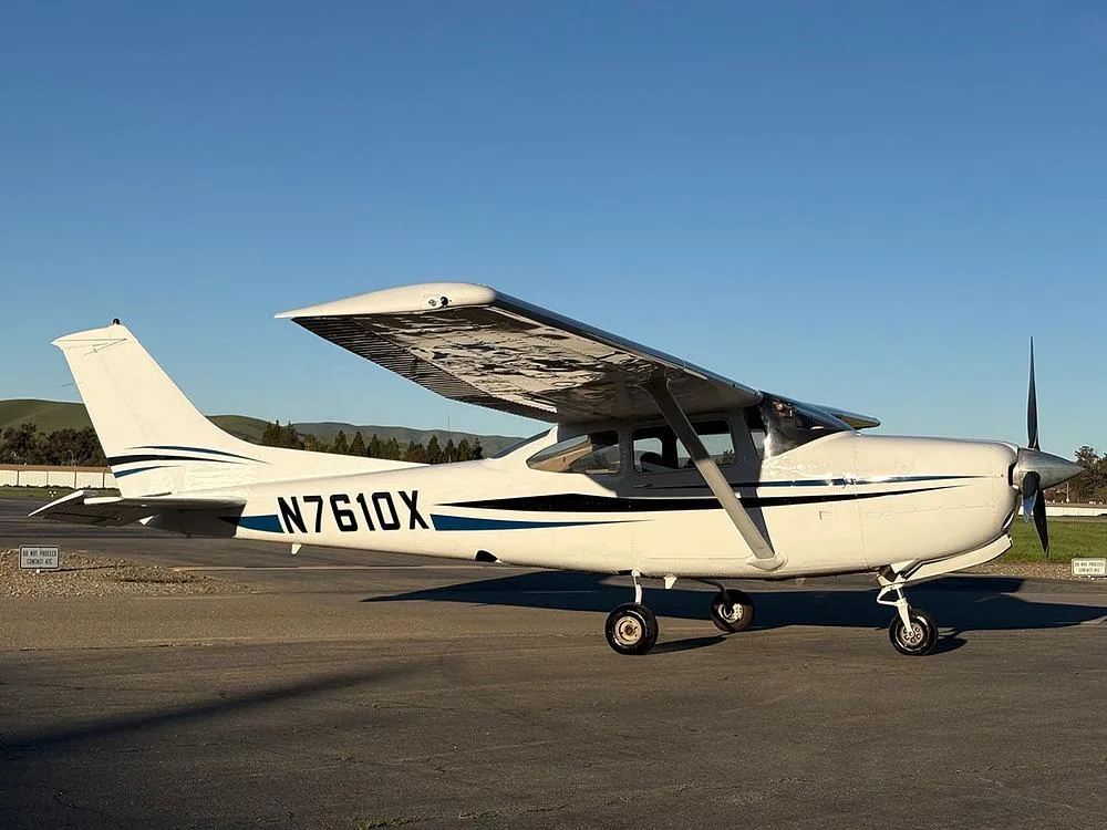A side view of a white Cessna 172 with blue and black stripes, registration N7610X, parked on an airfield.