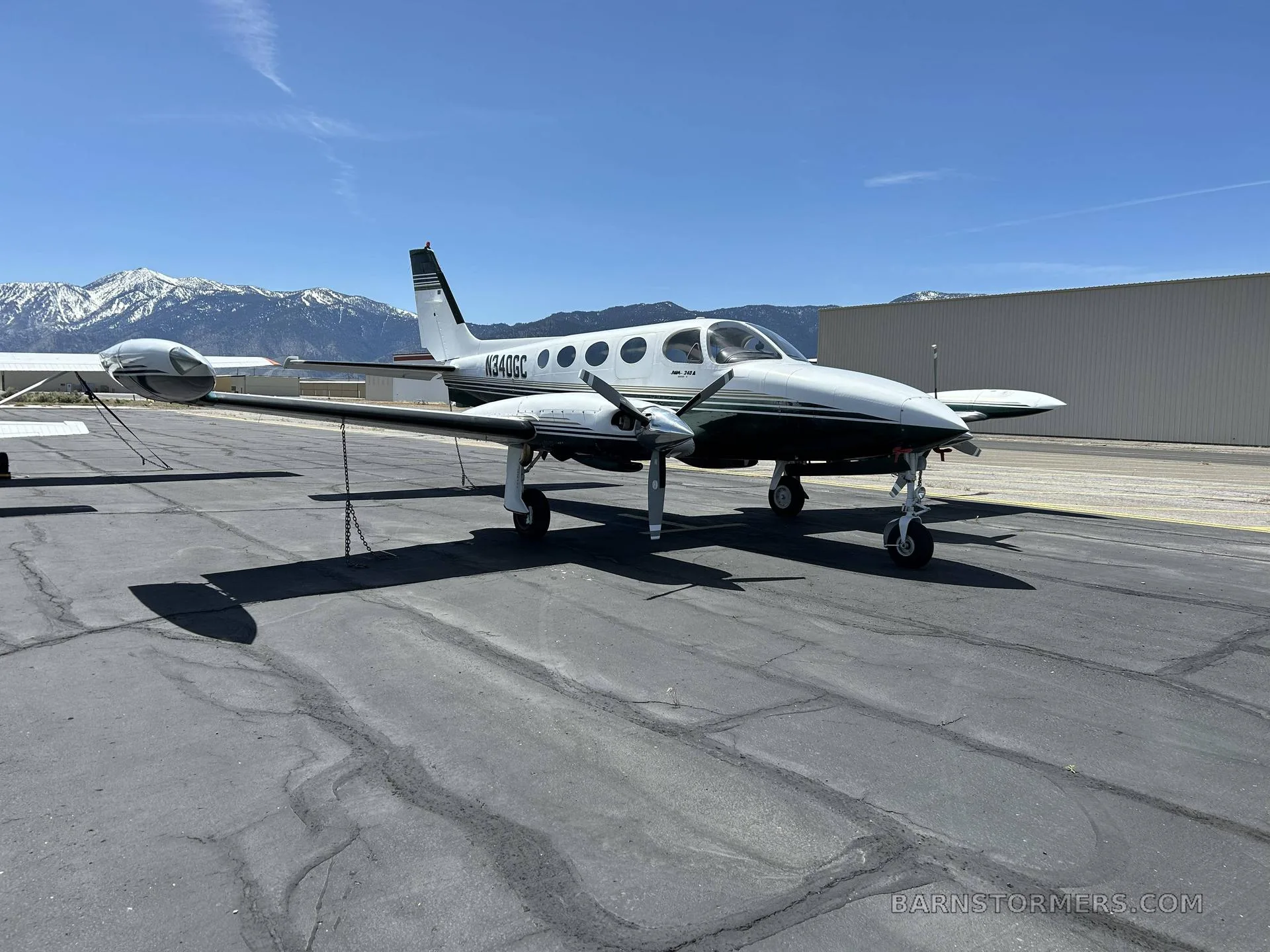 A white and dark green twin-engine propeller aircraft, registration N340GC, is parked on an asphalt ramp with snow-capped mountains in the background.