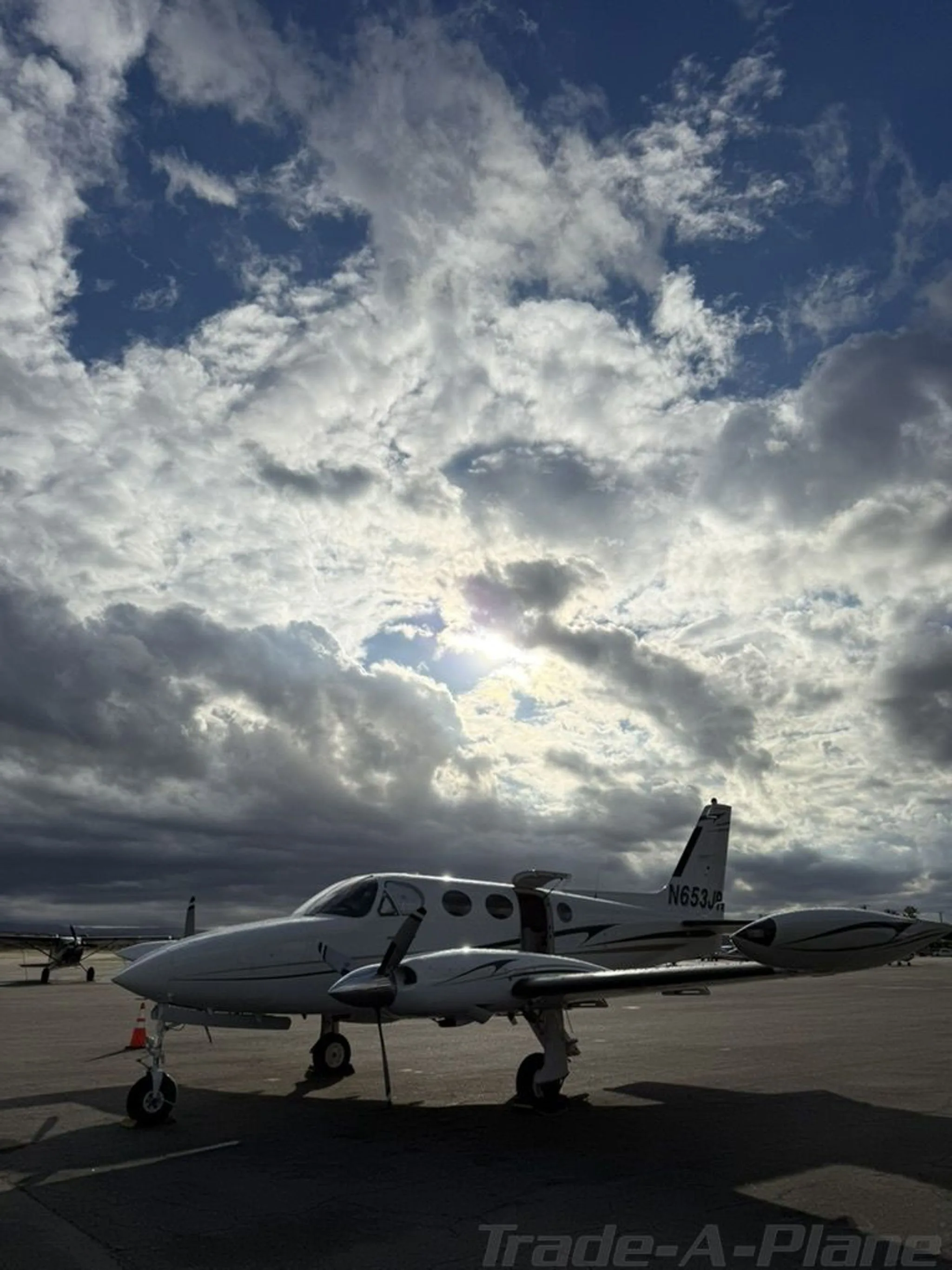 A white twin-engine aircraft, N653JR, is parked on a tarmac under a dramatic cloudy sky with the sun peeking through.
