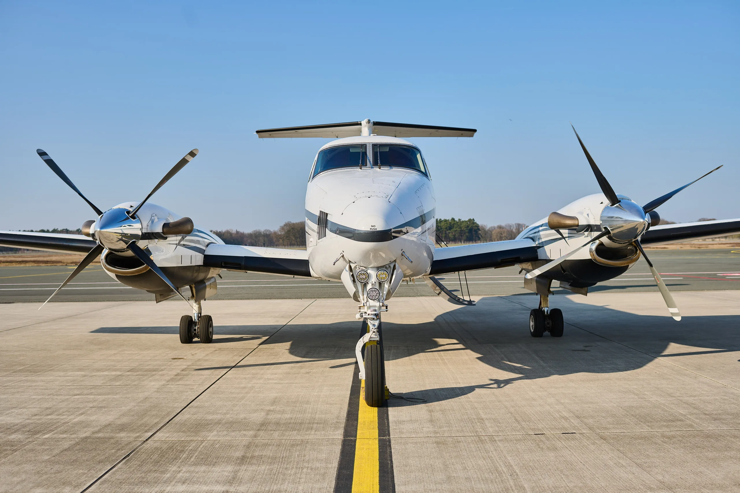 A head-on view of a white and black twin-turboprop aircraft, likely a King Air, parked on an airfield.