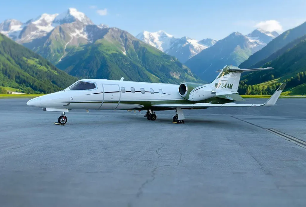 A white and green Learjet 31A (N784AM) is parked on an airfield with snow-capped mountains in the background.