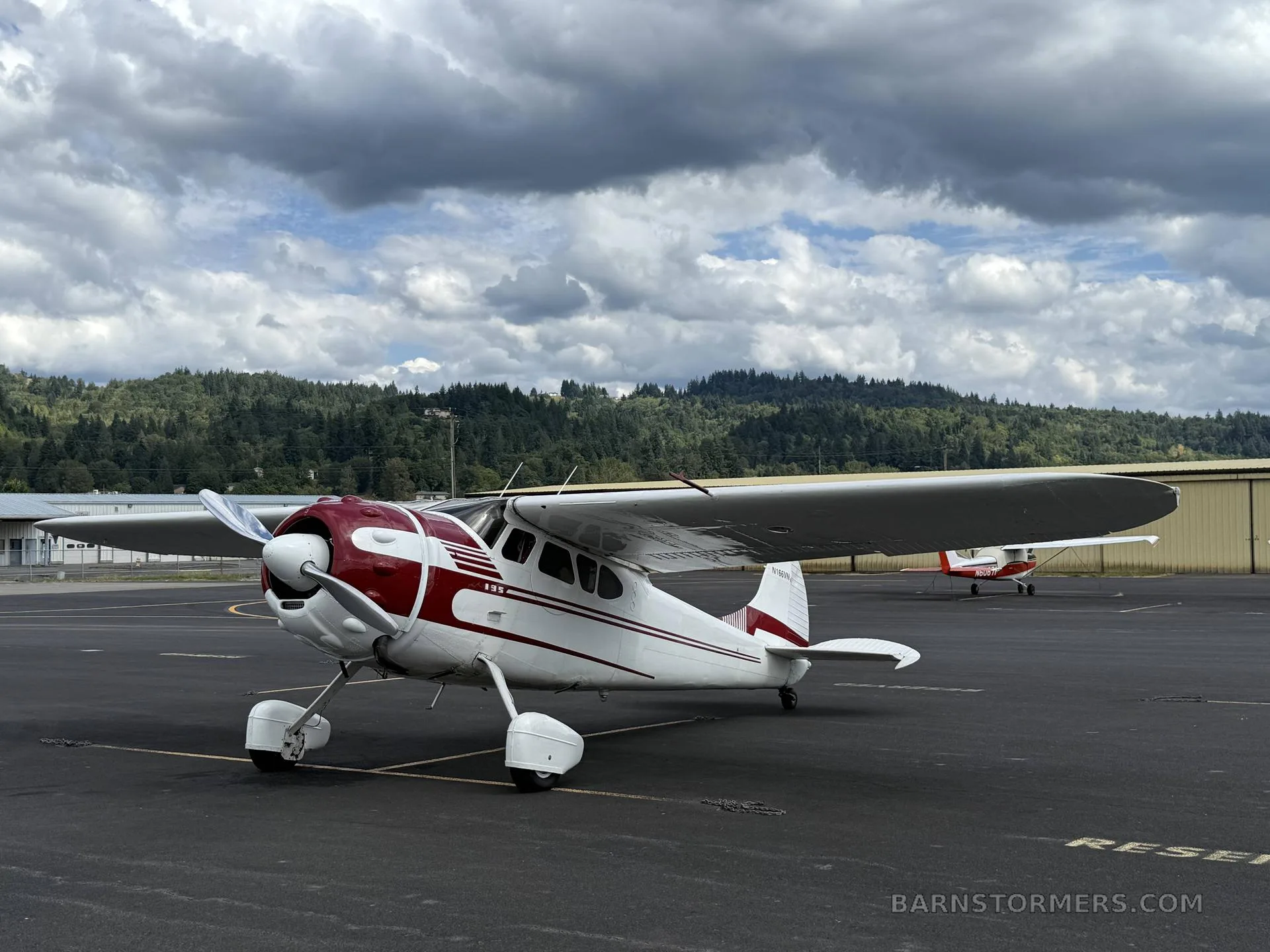 A white and red Cessna 180 or 185 on wheels, parked on an airfield tarmac under a cloudy sky with hills in the background.