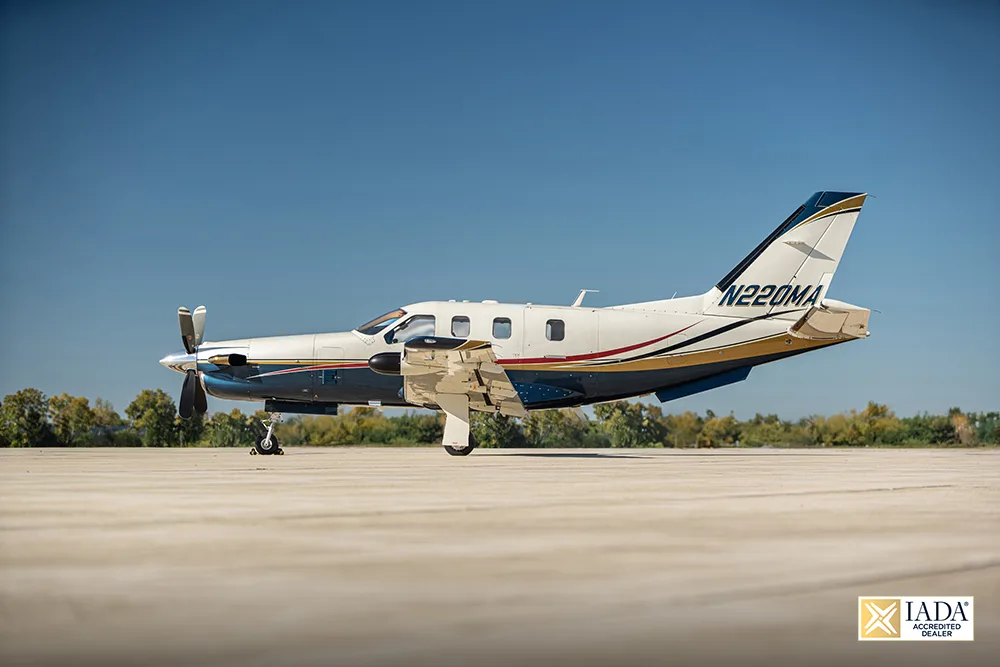 A white and blue turboprop aircraft with gold and red stripes, registration N220MA, parked on a tarmac under a clear blue sky.