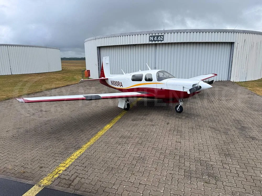 A white and red single-engine aircraft, N880RA, parked on a paved tarmac next to hangars under a cloudy sky.