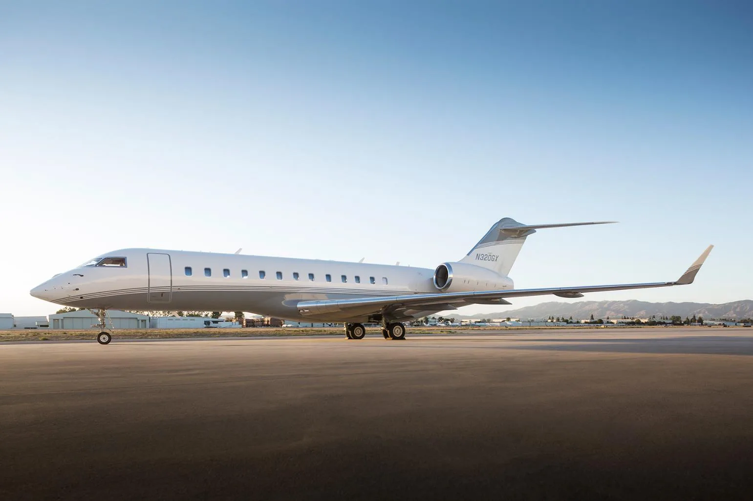 A white and grey business jet, tail number N320GX, is parked on an airfield with mountains in the background under a clear sky.