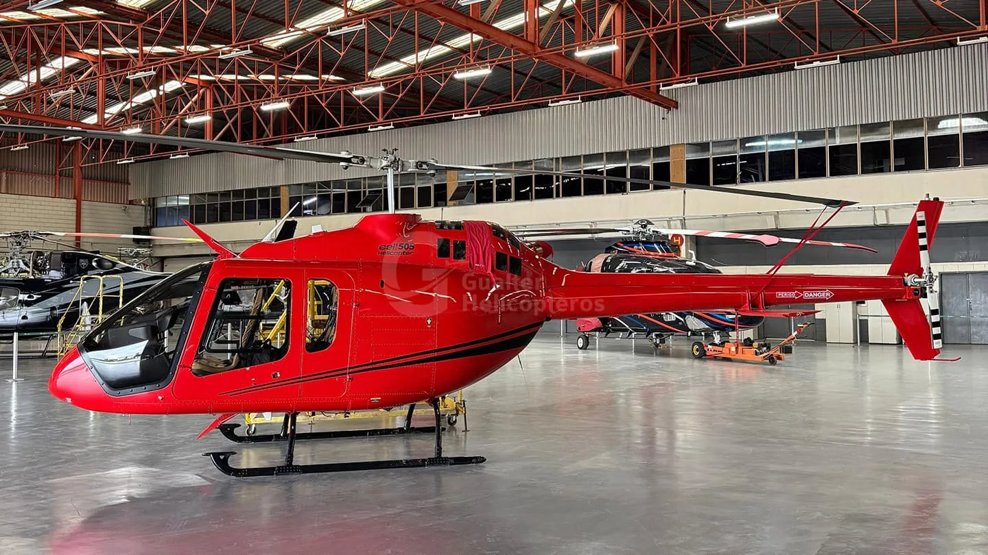 A bright red Bell 505 helicopter with black accents is parked inside a hangar, viewed from the side.