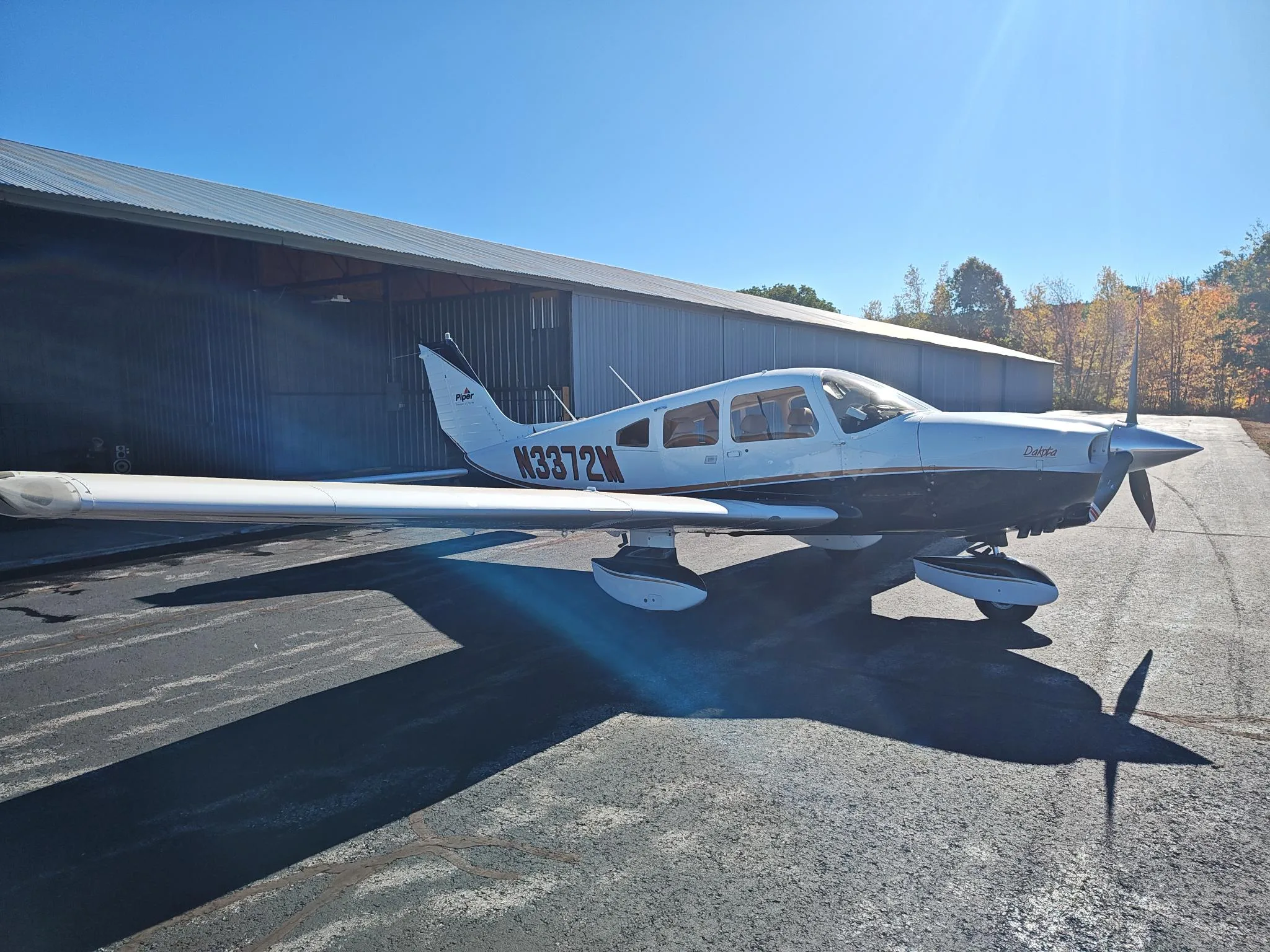 A white and dark-colored Piper Dakota aircraft with registration N3372M is parked on an asphalt apron next to a hangar on a sunny day with autumn trees in the background.