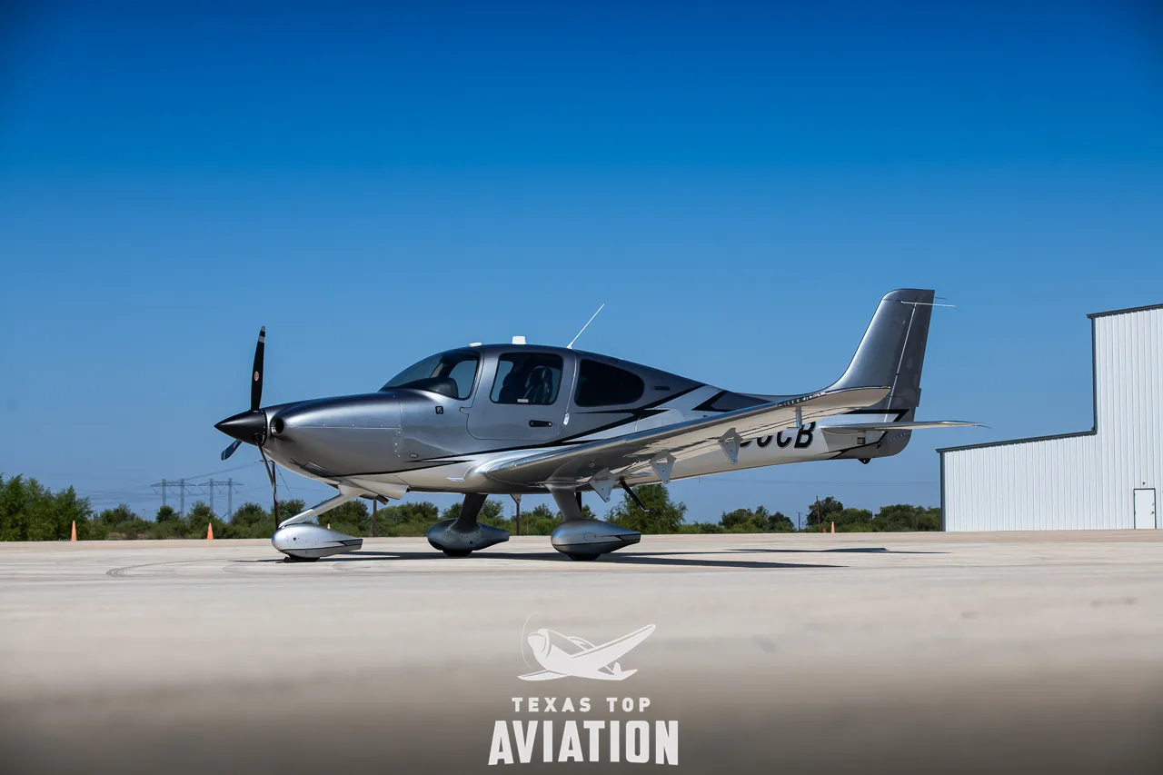 A side view of a silver and grey Cirrus SR22 aircraft with registration N886CB parked on an airfield.