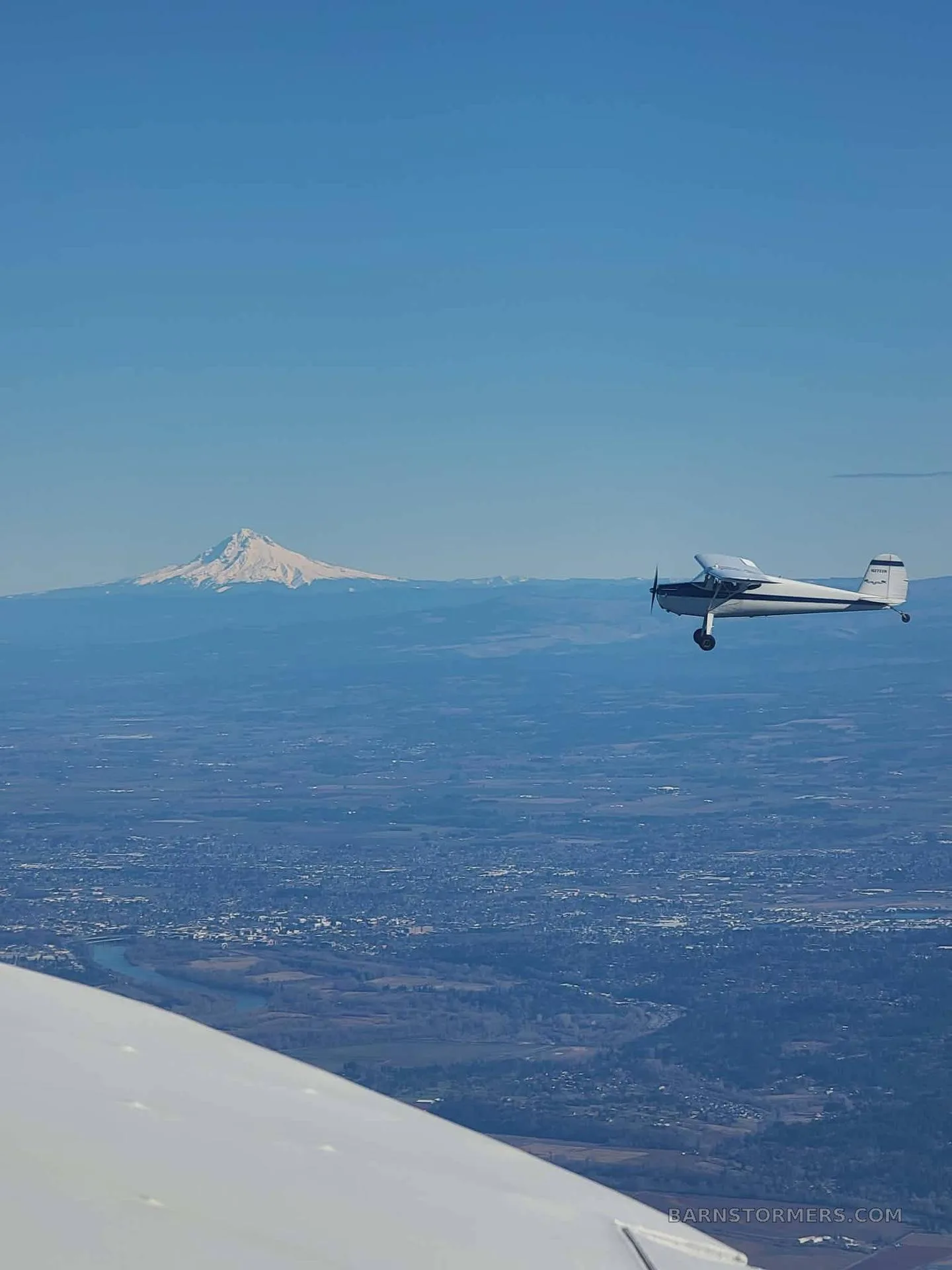 A white and blue taildragger aircraft flies over a city, with a snow-capped mountain in the distant background.
