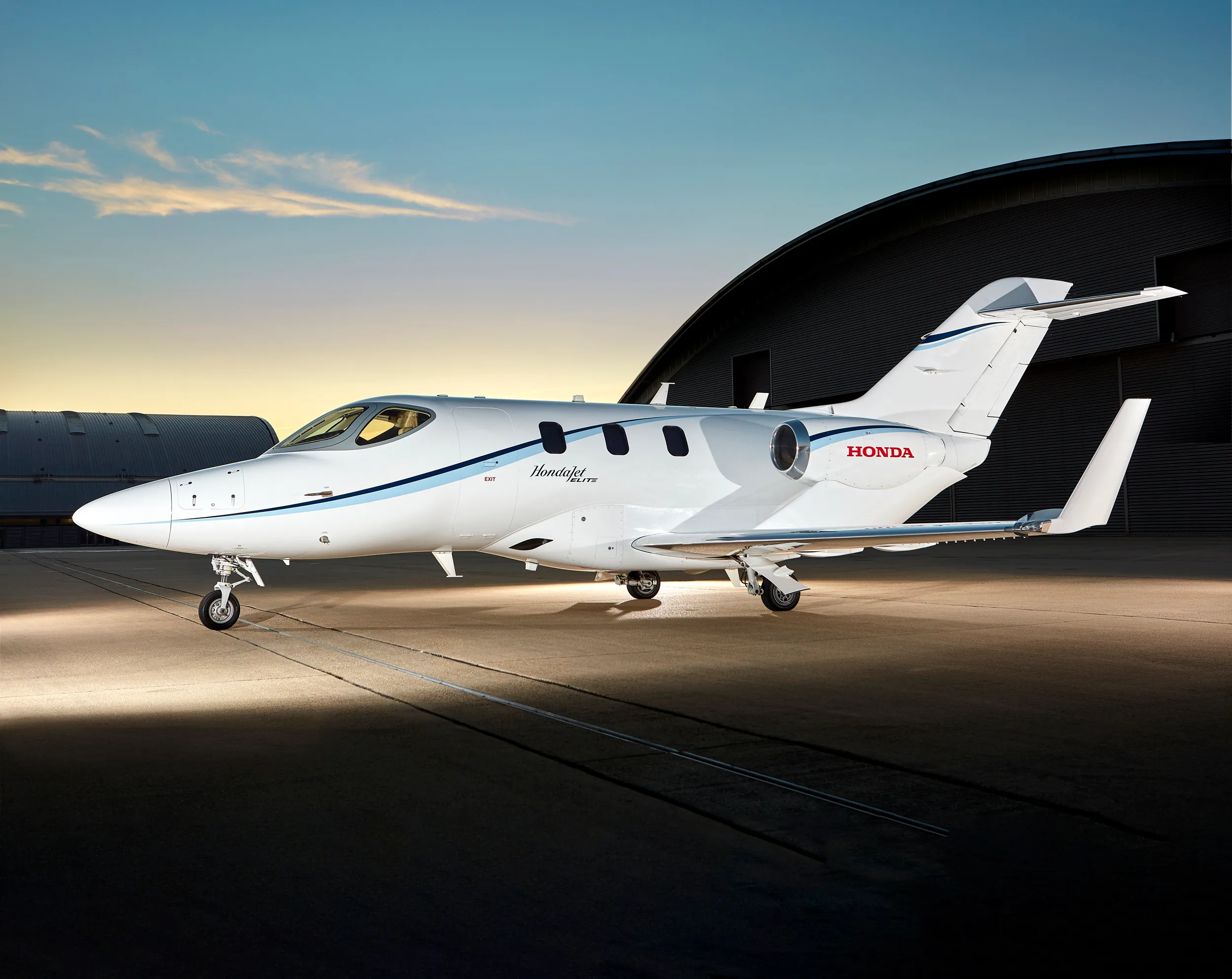 A white HondaJet Elite with blue stripes is parked on a tarmac in front of a hangar at sunset.
