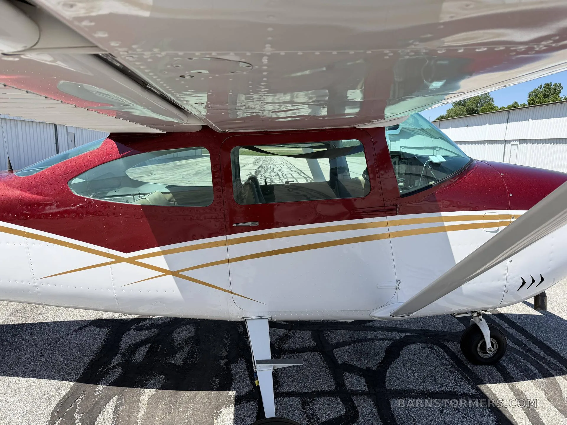A close-up side view of a red and white Cessna 182 aircraft with gold stripes on the tarmac.