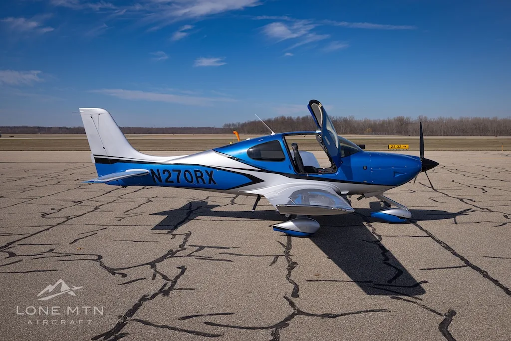 A side view of the blue and silver Cirrus SR20 aircraft, registration N270RX, with its doors open on an airport tarmac.