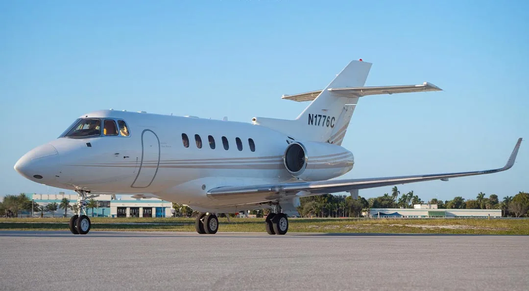 A white Hawker 800 series business jet with a light grey stripe is parked on an asphalt tarmac under a clear blue sky.